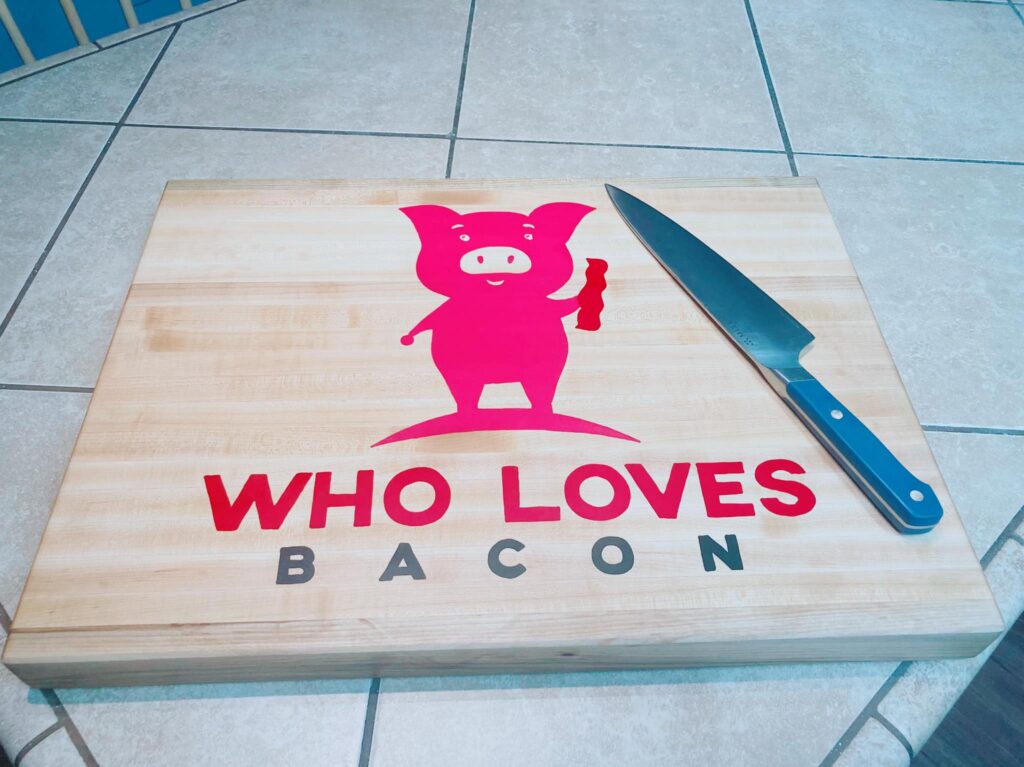 Cutting board with  butcher knife on it. 
Pink pig holding bacon
words WHO LOVES BACON